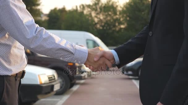 Dva obchodníci si potřásají rukama s auty na parkovišti v pozadí. Potřesení rukou mezi manažerem nebo dealerem a klientem. Potřesení mužských paží venku. Zavřít Zpomalený pohyb - Záběry, video