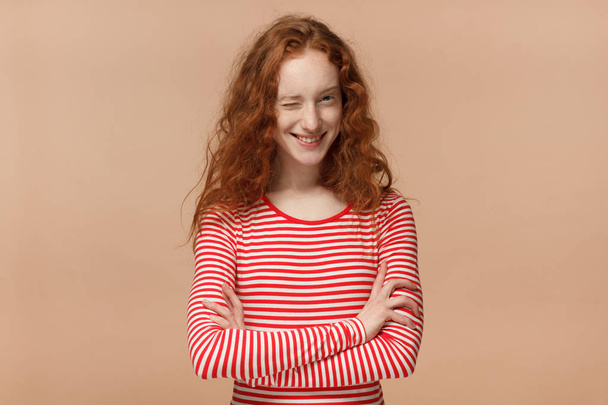 Pretty curly redhead girl in striped t-shirt with crossed arms smiling and winking, looking at camera  - Foto, afbeelding