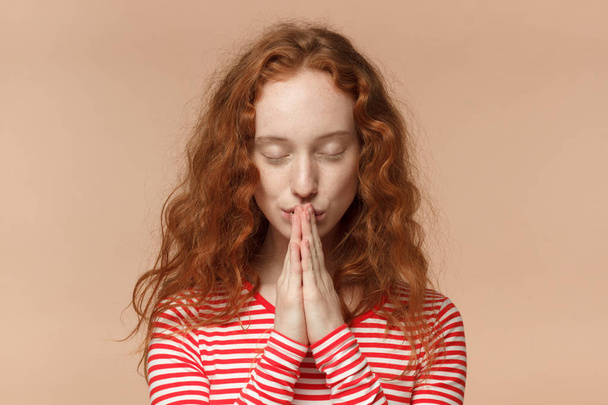 Closeup of attractive young redhead female in striped t-shirt putting hands together as if she is praying with closed eyes - Foto, Bild