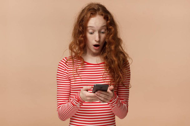 Portrait of attractive young redhead female, holding smartphone, looking scared and confused reading text message or e-mail - Fotó, kép