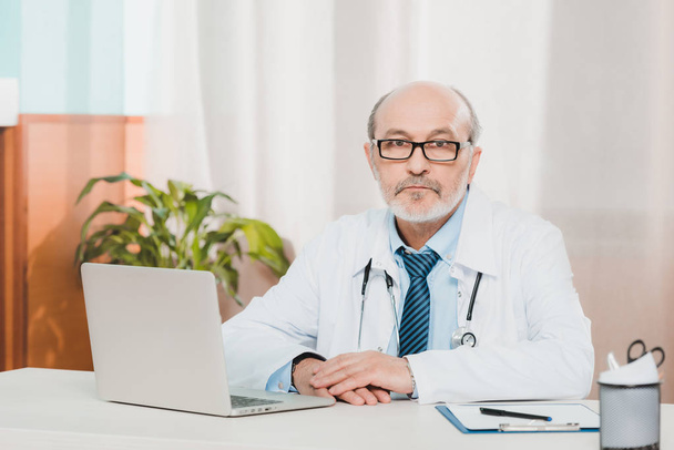 Porträt eines Oberarztes mit Brille, der am Arbeitsplatz mit Laptop in der Klinik in die Kamera blickt - Foto, Bild