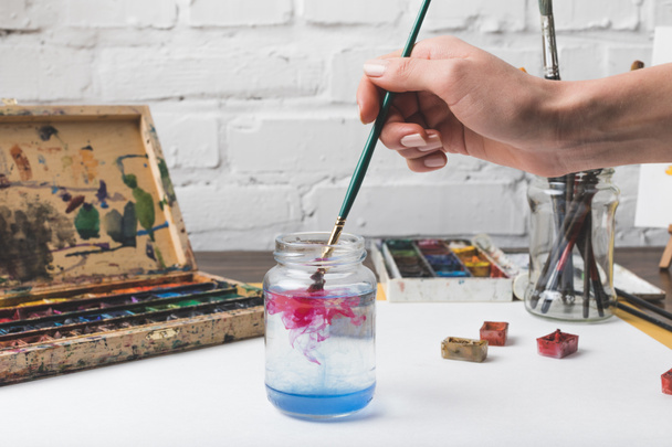 partial view of artist putting paint brush into glass jar with water at workplace - Photo, Image