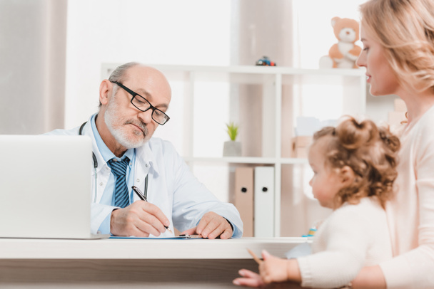 vanhempi lääkäri tekee muistiinpanoja, kun äiti ja pieni tytär istuu pöydässä lähellä klinikalla
 - Valokuva, kuva