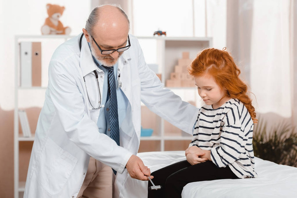 senior pediatrician in white coat examining little patient with reflex hammer in clinic - Photo, Image