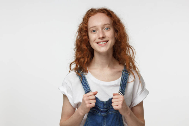 Indoor portrait of young beautiful redhead European female isolated on white background holding straps of jumpsuit in hands, showing inspired smile to her mate with relaxed and friendly look - Fotografie, Obrázek