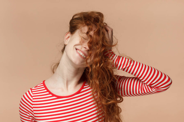 Closeup portrait of good-looking redhead teen girl isolated on peach background pressing loose curly hair to her head creating messy hairstyle looking extremely happy having fun with eyes closed - Fotó, kép