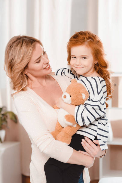 portrait of smiling mother holding little daughter with teddy bear - Photo, image