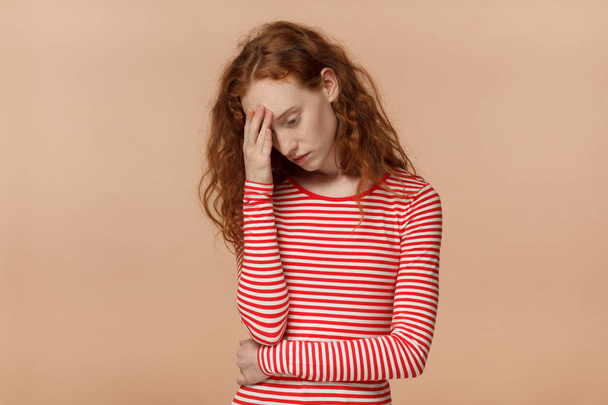 Studio photo of young attractive European lady with red curly hair isolated on peach background pressing palm to forehead looking worried, bored and exhausted with hard mind work or personal trouble - Foto, Bild