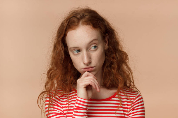 Closeup headshot of beautiful redhead woman isolated on peach background looking sidewards feeling distrust, hesitation, doubting given facts, uneasy to communicate and cooperate, showing discontent - Photo, image