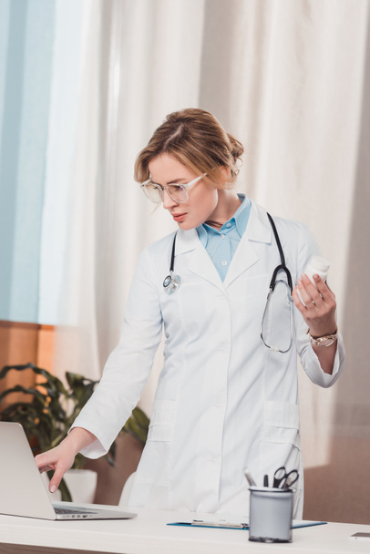 retrato de médico de casaco branco com medicamentos na mão usando laptop no local de trabalho na clínica
 - Foto, Imagem