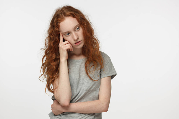 Tired and bored redhead female in gray t-shirt, gesturing with hand, solving relationship or health problem. Doubt concept.  - Photo, Image