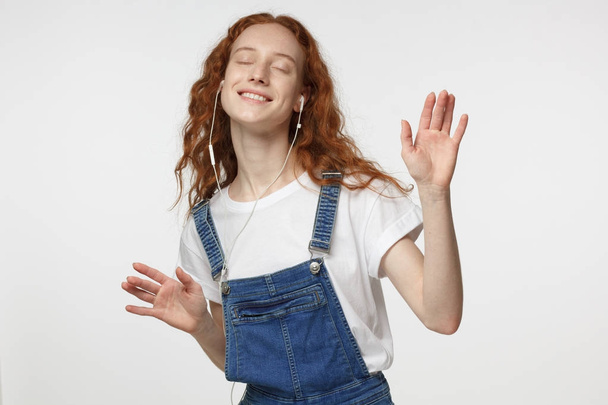 Young redhead female in denim jumpsuit in headphones listening to his favourite song, dancing, smiling with closed eyes - Valokuva, kuva