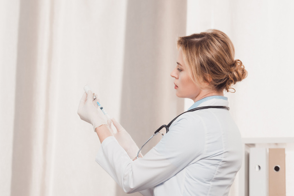 Zijaanzicht van doctor in de witte jas en medische handschoenen bedrijf spuit voor injectie in handen in kliniek - Foto, afbeelding