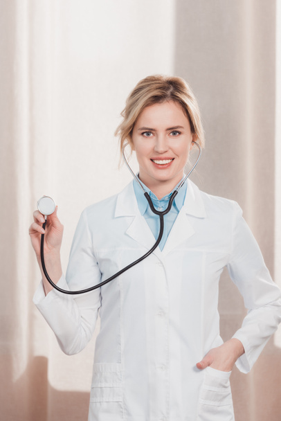 портрет усміхненої жінки-лікаря в білому пальто, що показує стетоскоп в руці
 - Фото, зображення