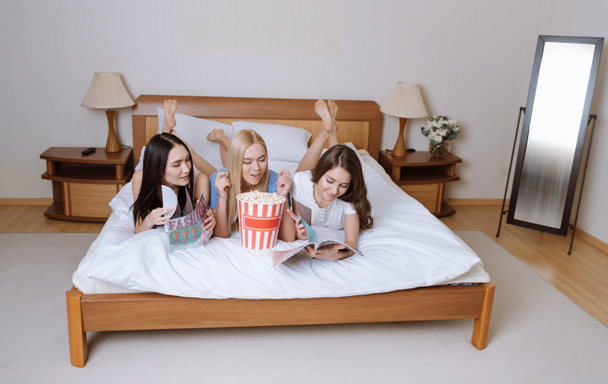 щасливі багатоетнічні дівчата лежать на ліжку з попкорном і читають журнали
 - Фото, зображення