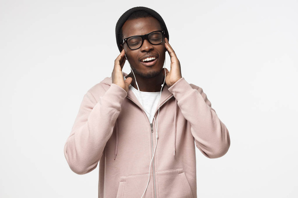 Portrait of happy young black man in pink hoodie, holding earphones with both hands; listening music wiht closed eyes; isolated on gray background - Photo, image