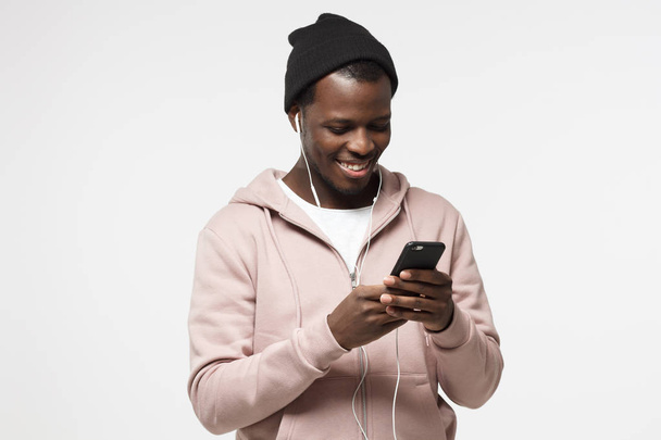 Young handsome black guy in pink hoodie and black hat, listening music with headphones and looking at screen of his smartphone - Foto, Imagen