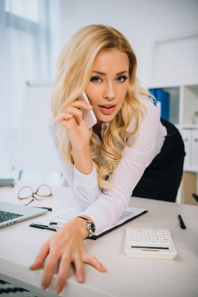 sexy businesswoman leaning on table and talking by smartphone - Photo, Image