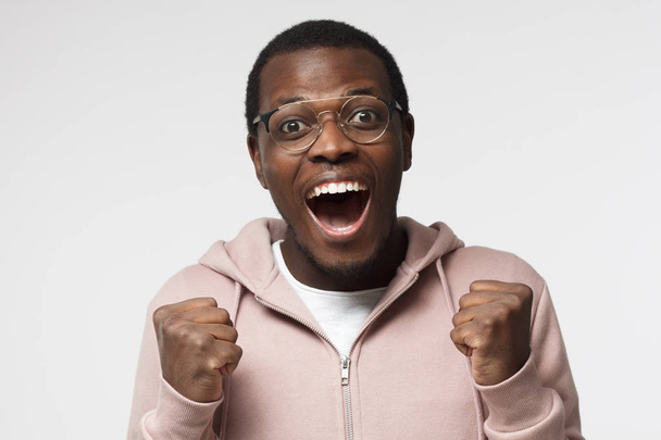Tiro horizontal de homem africano emocional isolado em fundo branco com óculos redondos na moda, apertando punhos e tendo aberto a boca em euforia alegre como se sentindo felicidade sobre a vitória
 - Foto, Imagem