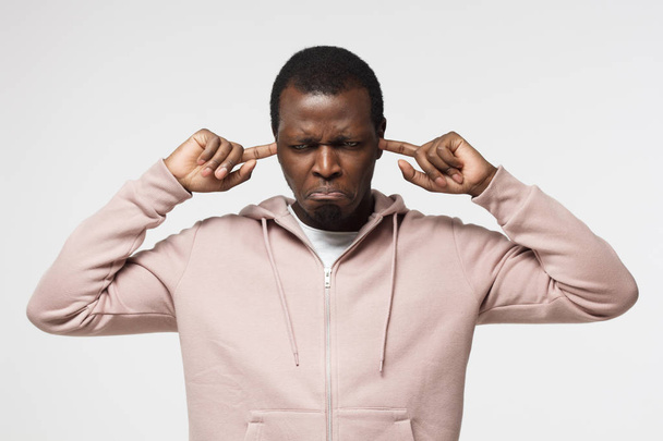 Horizontal shot of young African American male pictured isolated on white background stopping up his ears with sad disappointed face unwilling to hear unpleasant information or face problems - Foto, Imagem