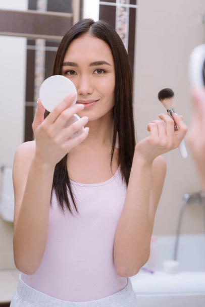 asiático chica holding base polvo y maquillaje cepillo en cuarto de baño
 - Foto, imagen