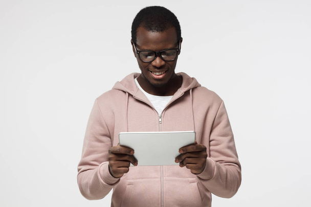 Studio closeup of young African American man pictured isolated on white background wearing eyeglasses looking attentively to screen of tablet he is holding, smiling happily, looking involved - Foto, Imagem