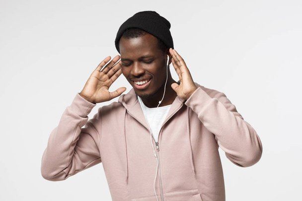 Indoor picture of handsome African American guy isolated on white background wearing hat and hoodie rising up hands while enjoying audio track through white earphones, smiling with eyes closed - Photo, image