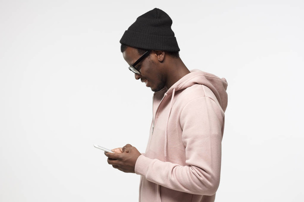 Side portrait of handsome African American guy in pink hoodie and black hat isolated on white background looking attentively at screen of smartphone while exchanging messages or surfing Internet - Photo, image