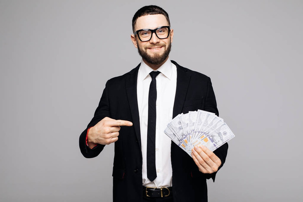 Portret mężczyzny udany uśmiechający się w garnitur i okulary, wskazując palcem na kilka banknotów pieniądze na białym tle nad szarym tle - Zdjęcie, obraz
