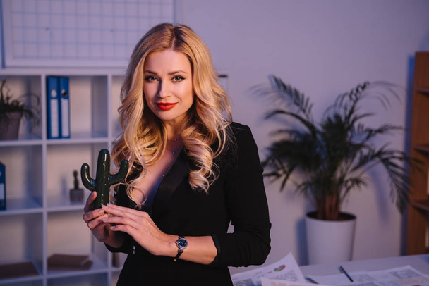smiling sexy woman holding artificial cactus in office and looking at camera - Фото, зображення