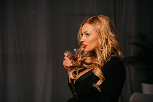 side view of attractive sexy blonde woman drinking whiskey in office at evening - Foto, afbeelding