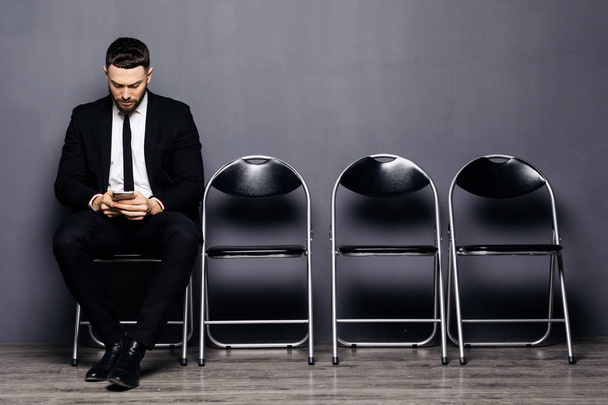 Готов к интервью. Красивый мужчина в формальной одежде держит телефон и использует его, сидя на стуле в комнате ожидания
 - Фото, изображение