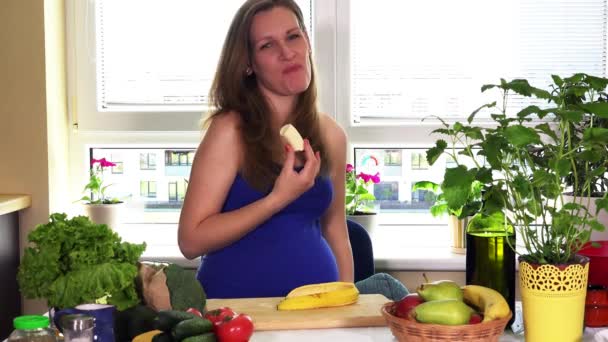 Pregnancy time nutrition. Pregnant woman eat banana sitting near kitchen table - Filmati, video