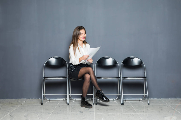 Jeune femme d'affaires ennuyée tenant du papier et regardant loin tout en étant assis sur une chaise sur fond gris
 - Photo, image