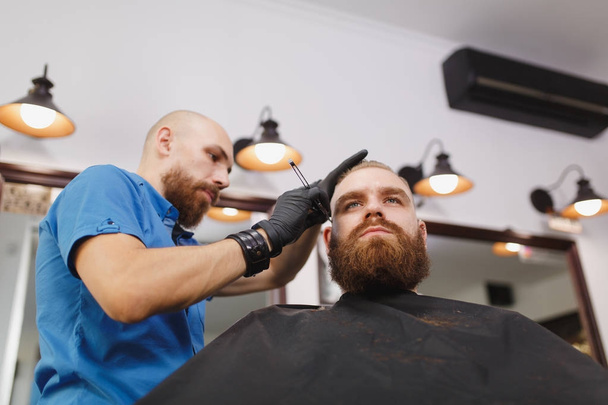 Мужчина профессиональный парикмахер, обслуживающий клиента, бреющий толстую большую бороду бритвой. Рыжий красивый стильный молодой человек с короткой стрижкой, черная накидка. Светло-белая парикмахерская
 - Фото, изображение