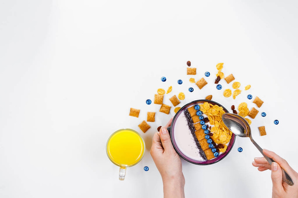 Breakfast, female hands holding a spoon over a bowl with homemade yogurt and cornflakes, and fresh blueberries, a glass of orange juice on a white background, top view, flat lay. Concept of healthy fo - Valokuva, kuva