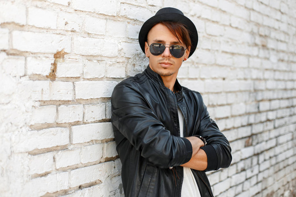 Szép elegáns fiatal férfi, napszemüveg, divatos kalap és fekete bőrkabát jelentő tégla fal közelében - Fotó, kép