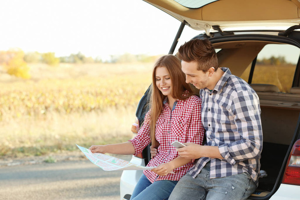 Beautiful young couple sitting with map in car trunk - Foto, imagen