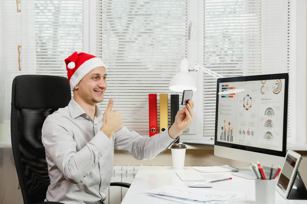 Красивый улыбающийся бизнесмен в рубашке и красной рождественской шляпе сидит
 - Фото, изображение