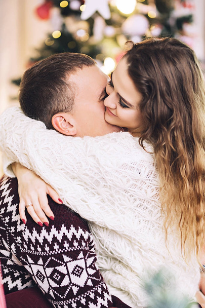Happy couple dressed in the sweaters is kissing and hugging near the christmas tree, cozy atmosphere and love, home and family, white caucasian couple, married people - Foto, imagen