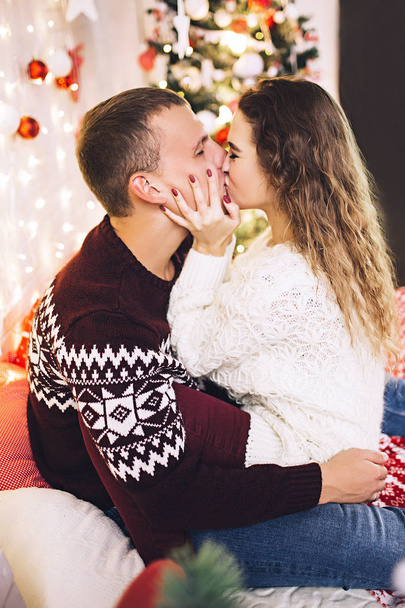 Happy couple dressed in the sweaters is kissing and hugging near the christmas tree, cozy atmosphere and love, home and family, white caucasian couple, married people - Photo, Image