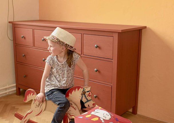Schattig klein meisje schommels op schommelpaard. Klein meisje draagt strohoed. Klein meisje speelt in de woonkamer. - Foto, afbeelding