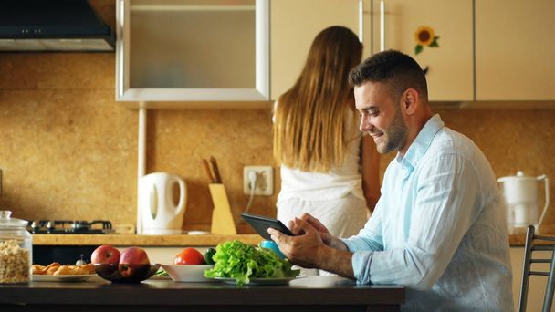Attractive couple chatting in the kitchen early morning. Handsome man using tablet while his girlfriend cooking - Foto, Imagen
