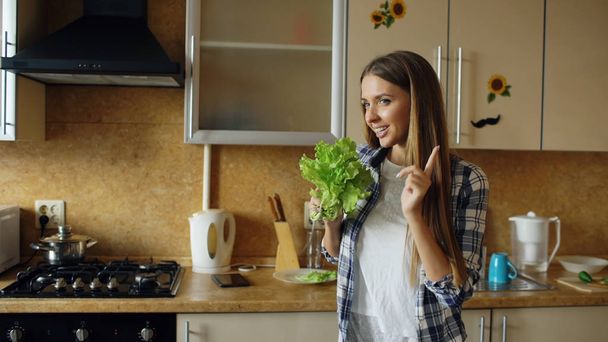 Cheerful young funny woman dancing and singing with lettuce microphone while cooking breakfast - Fotografie, Obrázek