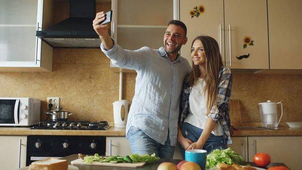 Young happy couple taking selfie picture while cooking breakfast in the kitchen at home - Фото, изображение