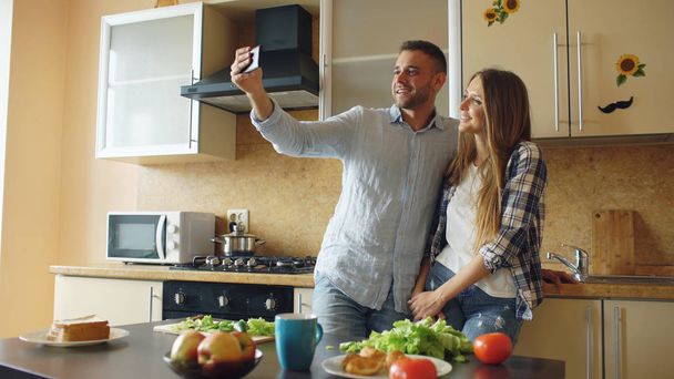 Young happy couple taking selfie picture while cooking breakfast in the kitchen at home - Foto, afbeelding