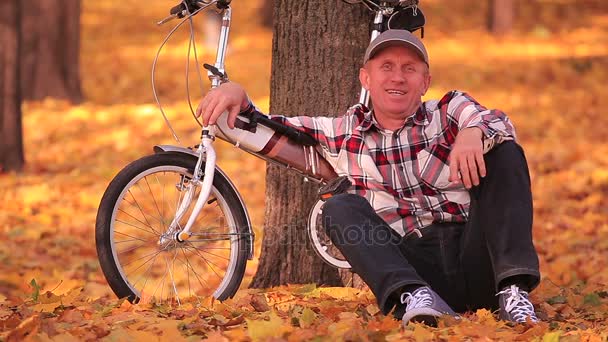 Dorosły człowiek siedzieć w pobliżu mały rower w jesienny park z drzewami żółty - Materiał filmowy, wideo