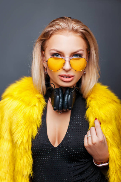 Модная стильная молодая женщина с наушниками в желтых очках моды и стильной желтой шубой на сером фоне
 - Фото, изображение