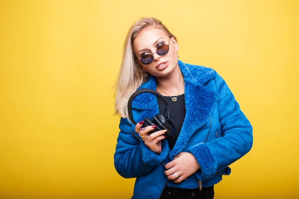 Young girl in round vintage sunglasses with headphones in a blue stylish jacket on a yellow background - Fotó, kép