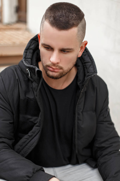 Retrato de um jovem com um cabelo curto em uma jaqueta de inverno quente senta-se nas escadas
 - Foto, Imagem